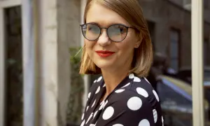 Kobieta w okularach i sukience w grochy, uśmiecha się do aparatu.