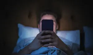 Mężczyzna leżący w łóżku i patrzący na ekran telefonu w ciemności.
