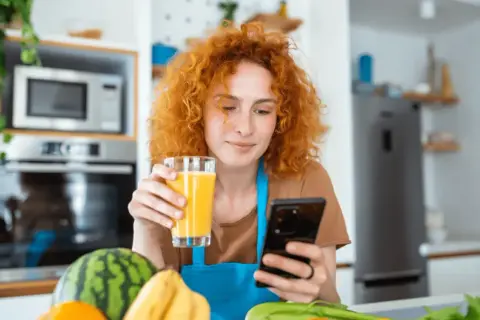 Rudowłosa kobieta z sokiem w dłoni szuka w telefonie informacji, co jeść na poprawę wzroku. Na blacie owoce i warzywa.