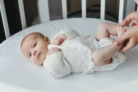 Noworodek w białej koszulce leży w łóżeczku. Dorośli delikatnie dotykają jego stópek. Tak jak widzi noworodek, świat jest rozmazany, ale pełen miłości.