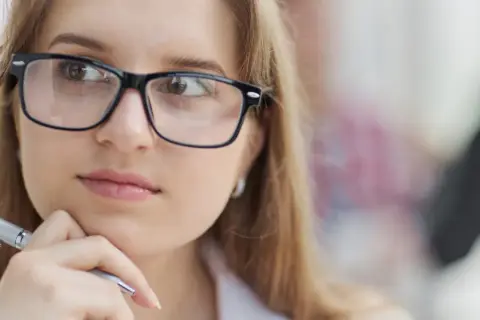Młoda kobieta w okularach, z długopisem w dłoni, zamyślona. Jej słaby wzrok nie przeszkadza jej w pracy.