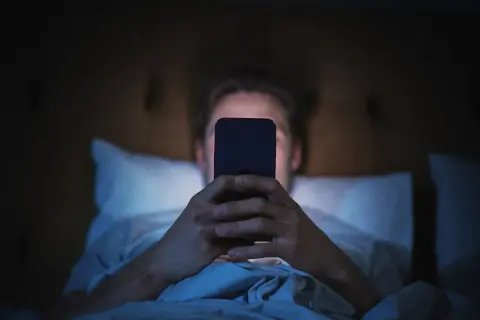 Mężczyzna leżący w łóżku i patrzący na ekran telefonu w ciemności.