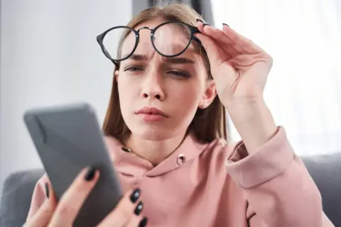 Zmarszczona kobieta w okularach trzyma telefon, szukając domowych sposobów na mroczki przed oczami.