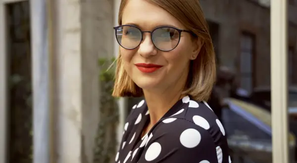 Kobieta w okularach i sukience w grochy, uśmiecha się do aparatu.