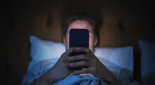 Mężczyzna leżący w łóżku i patrzący na ekran telefonu w ciemności.