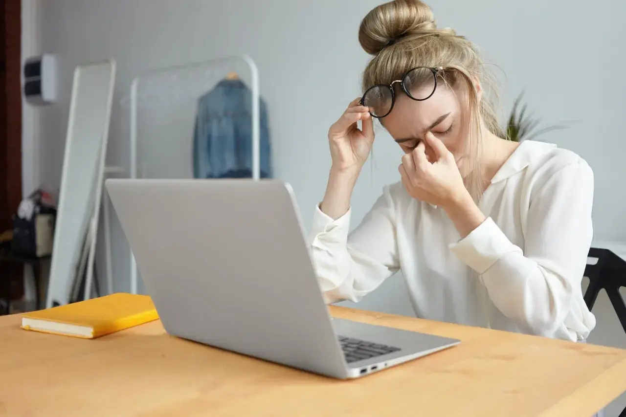 Kobieta w okularach masuje zmęczone oczy przy laptopie.