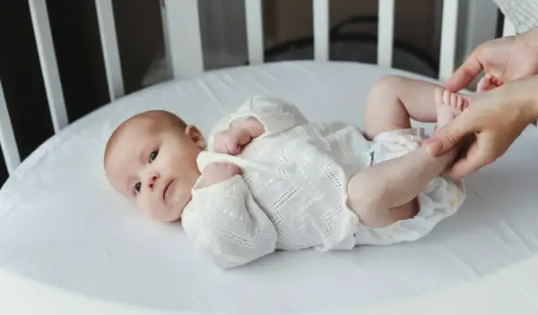 Noworodek w białej koszulce leży w łóżeczku. Dorośli delikatnie dotykają jego stópek. Tak jak widzi noworodek, świat jest rozmazany, ale pełen miłości.